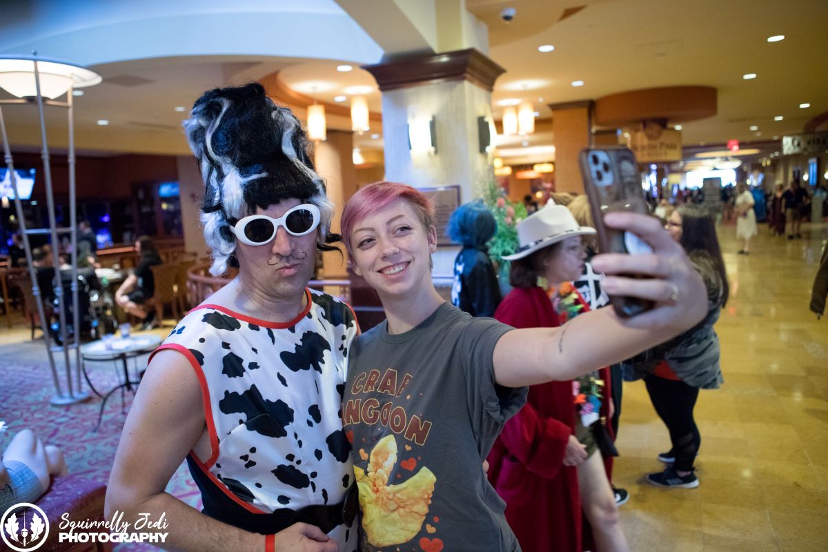 A con goer with pink short hair and a shirt that says "crab rangoon" smiles for a selfie photo with phone outstretched with a cosplayer in a black and white beehive wig, white sunglasses and cow print top.