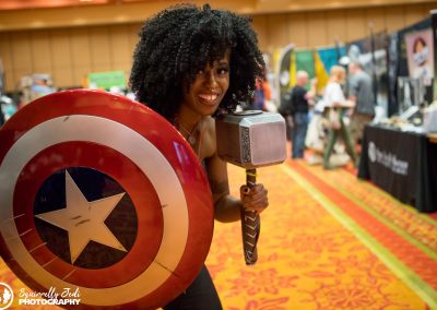 A female Captain America holds forward her shield with her right hand and holds Mjölnir up by her face in her left hand.