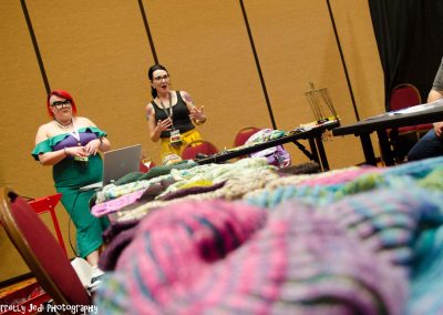 Two teachers stand at the front of the class speaking about knitting. There is a close up shot of knitted projects in the foreground.
