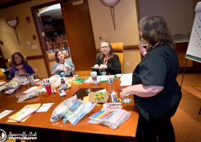 A teacher stands at the head of a conference table speaking to students as they do a craft with paper. There is paper and supplies on the table.