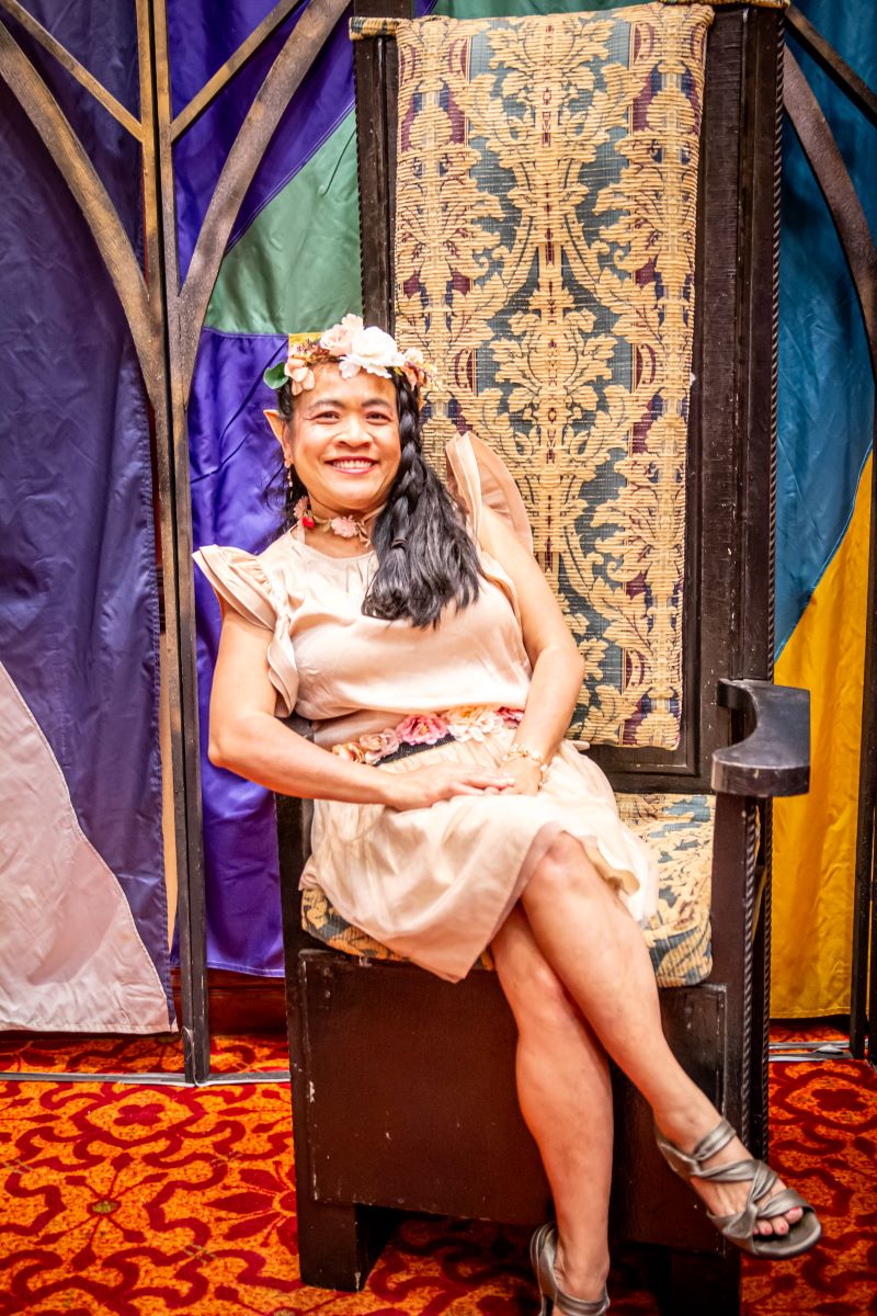 IMG_1094 A woman dressed as an elf with a flower crown and pink dress sits in a throne chair in the club area of Soonercon.